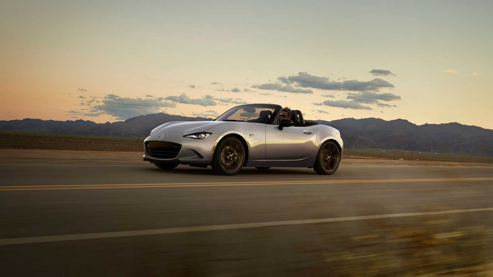 Dynamic front 3/4 shot of a silver 2024 Mazda MX-5 driving on a country road with mountains in the background.